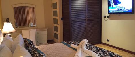 Una habitación de hotel con cama, televisión en la pared, armario y escritorio con lámpara.