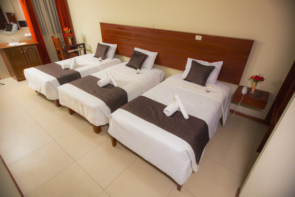 A hotel room with three neatly made beds, a wooden headboard, a small desk with a chair, and a nightstand with a phone and a vase with flowers.