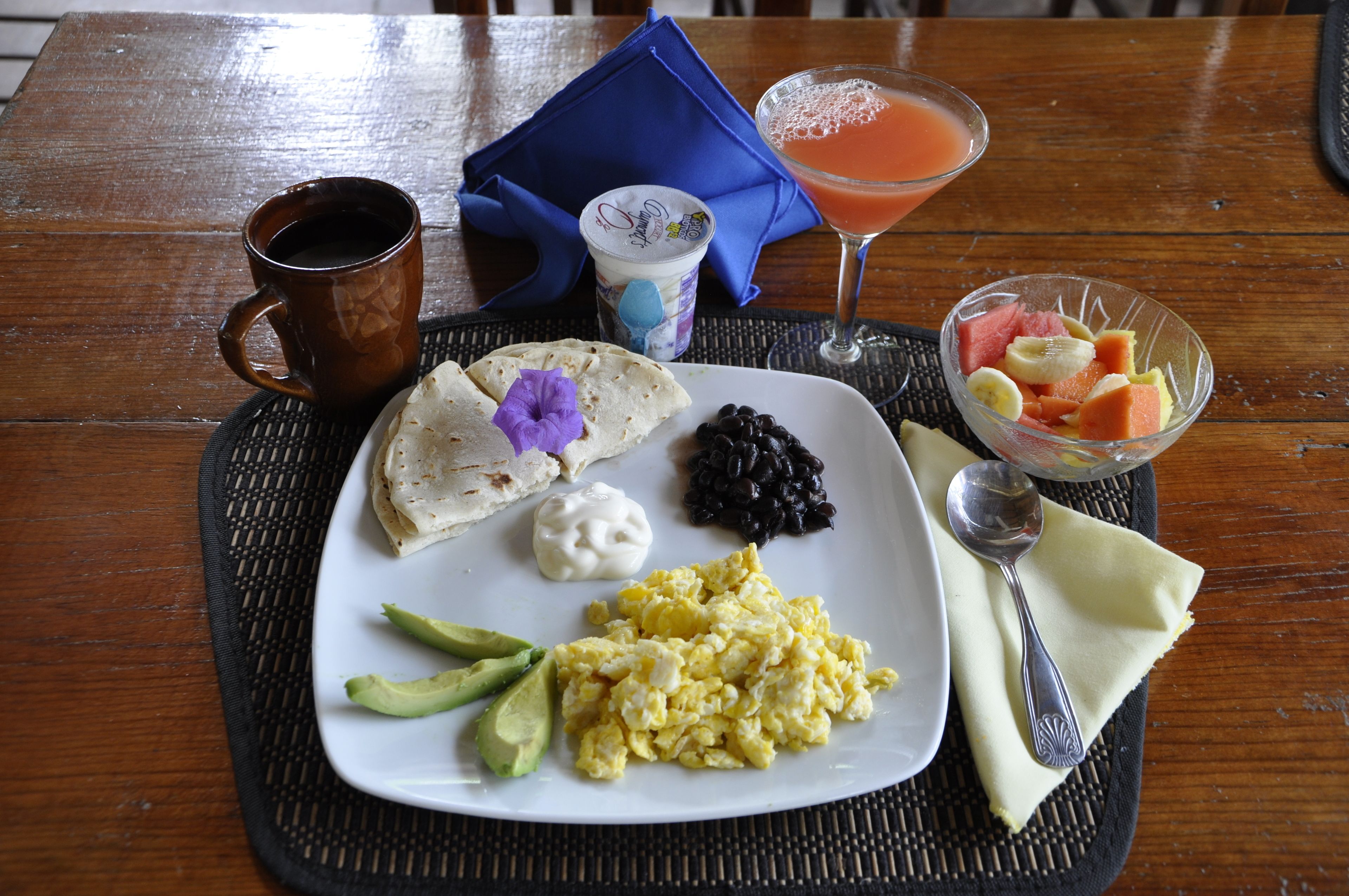 Un plato con huevos revueltos, frijoles negros, rebanadas de aguacate y una tortilla decorada con una flor morada, acompañado de una taza de café y un vaso de jugo de naranja sobre una mesa de madera.
