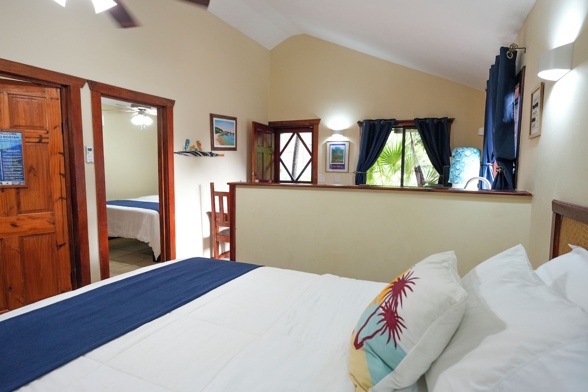 A hotel room with a bed, a wooden door, a window with curtains, and a wall-mounted light fixture.