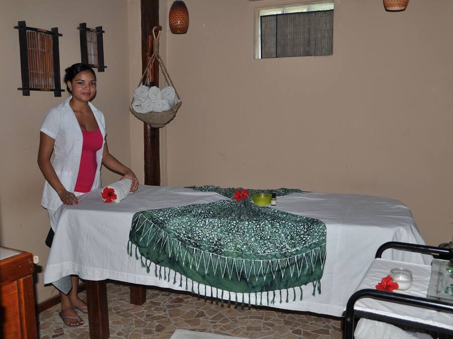 A room with a bed covered in a white sheet and a green floral blanket, a small table with a candle and a glass, and a hanging basket with towels.