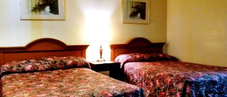 Habitación de hotel con dos camas individuales, una mesita de noche con lámpara y dos cuadros en la pared.