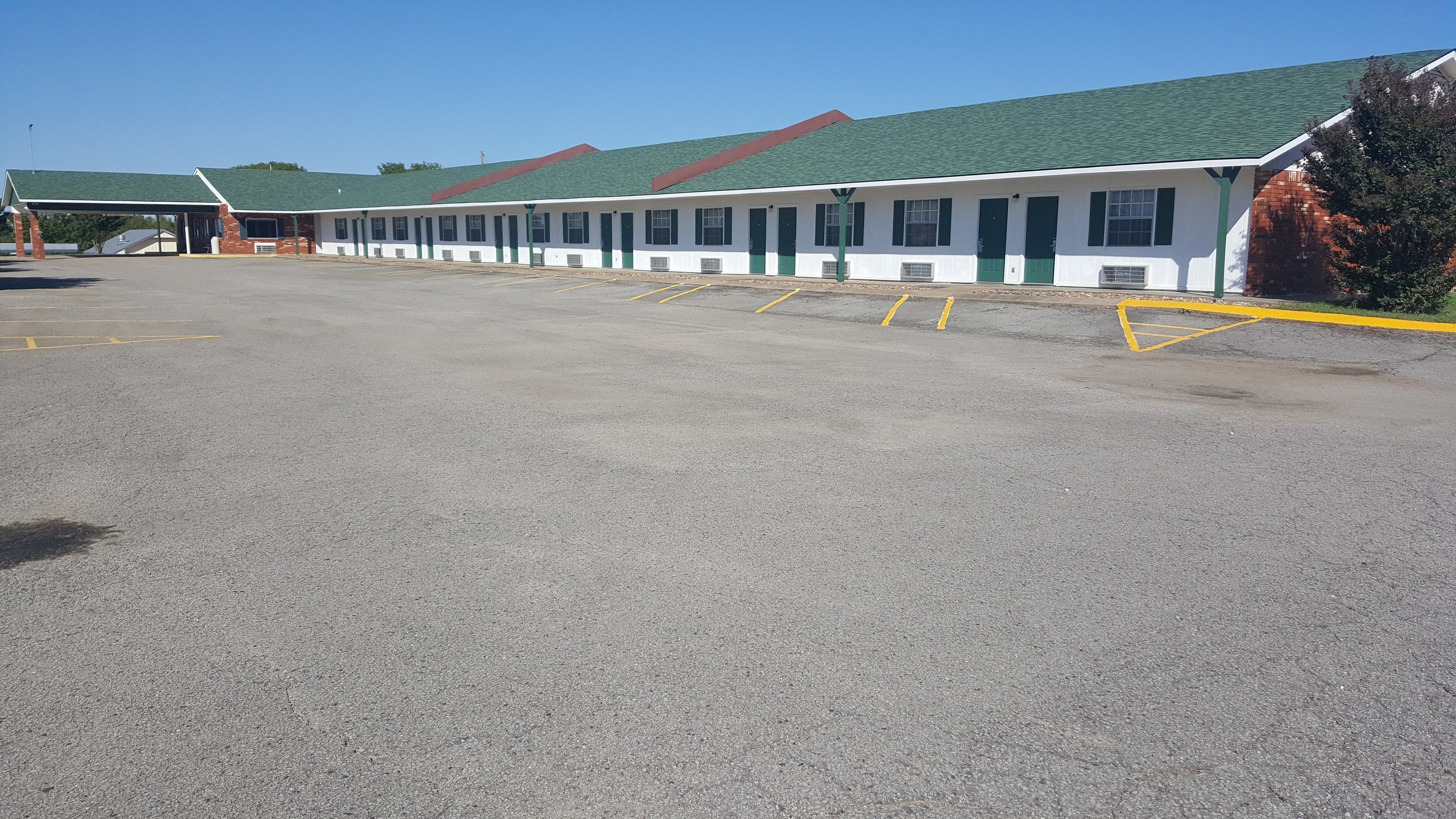 A motel with a green roof and white exterior walls, featuring a row of rooms with green doors and windows, and a parking lot with yellow lines.