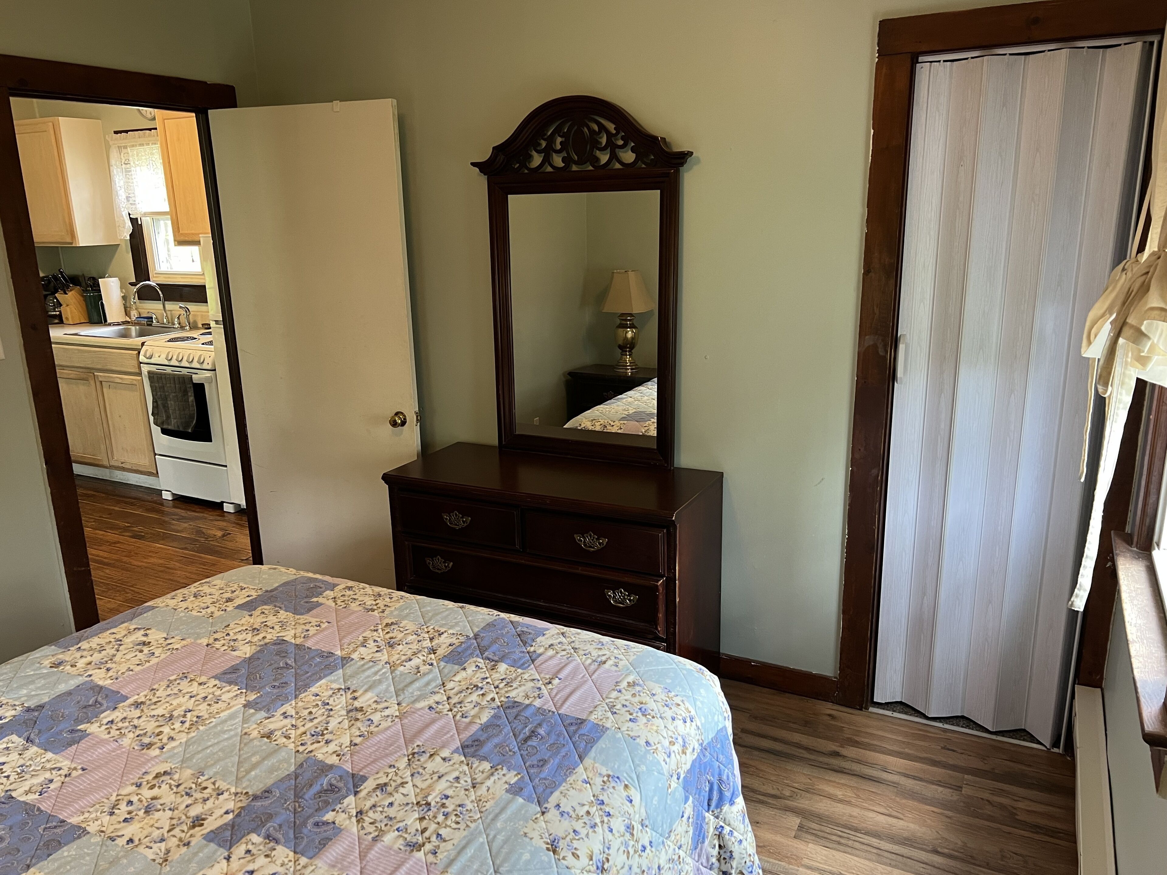 Un dormitorio con cama, cómoda, espejo y puerta que da a una cocina.