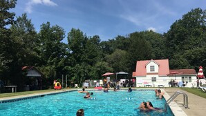 A swimming pool with people in it, surrounded by trees and a building with a red roof. - Echo Valley Cottages (East Stroudsburg)