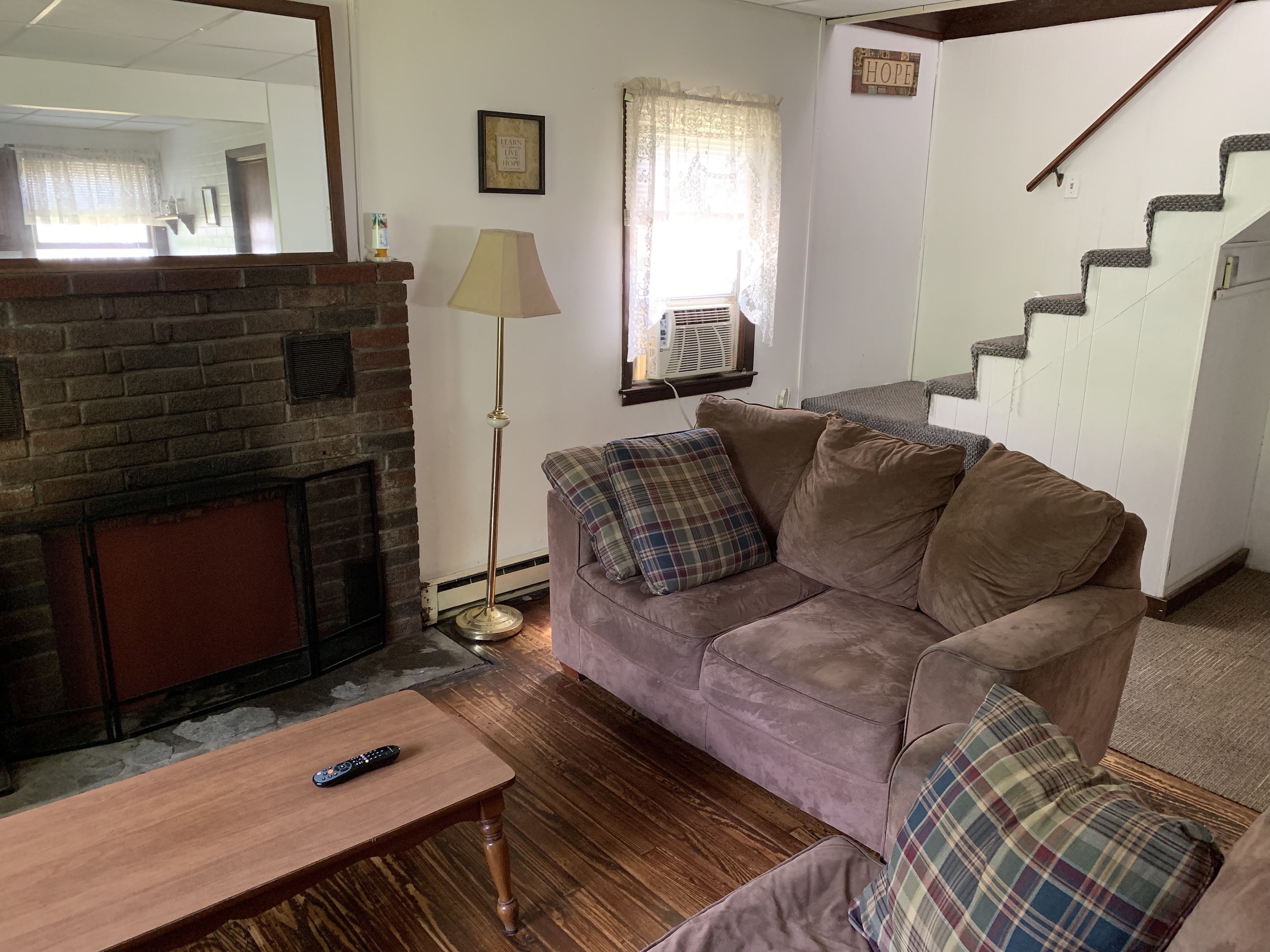 A cozy living room with a brick fireplace, a sofa with plaid cushions, a wooden coffee table, and a staircase.