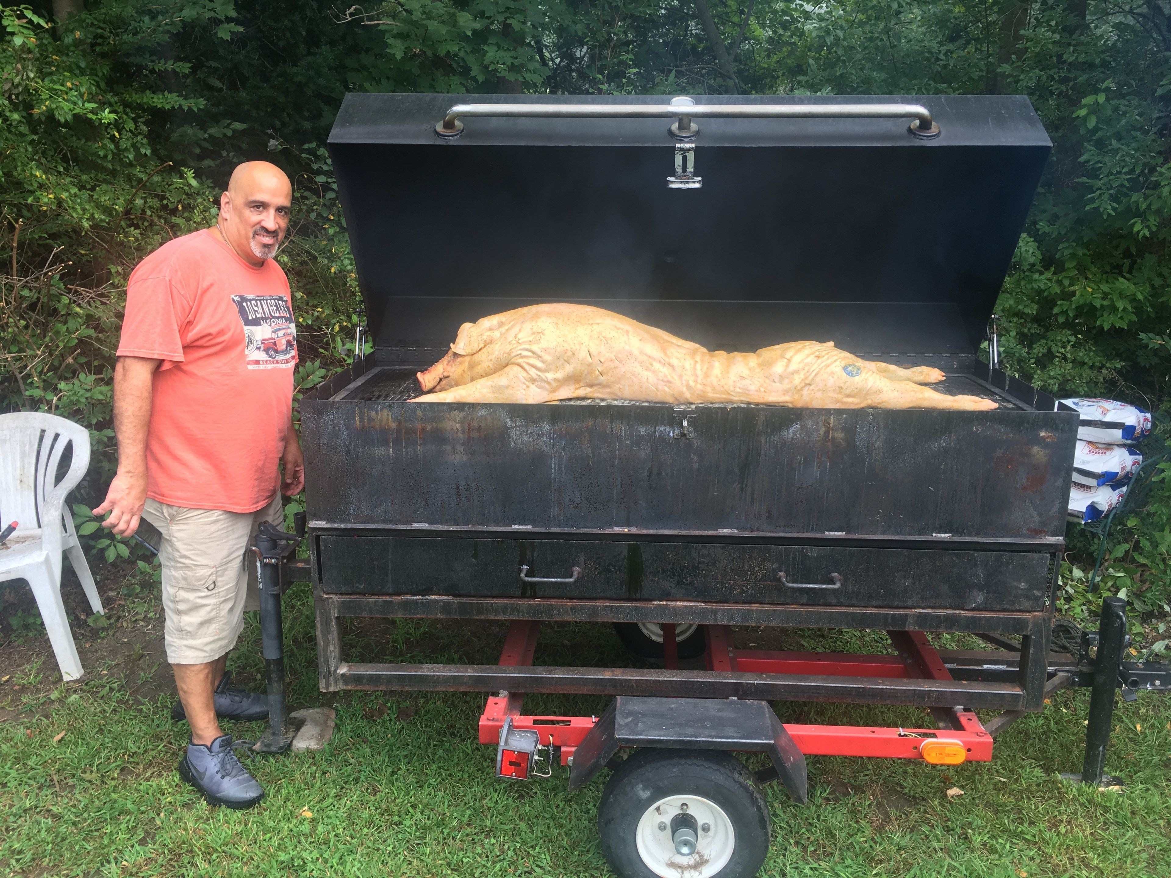 Un grand barbecue extérieur sur lequel est posé un gros morceau de viande, un homme qui se tient à côté, et une chaise en plastique blanche à proximité.