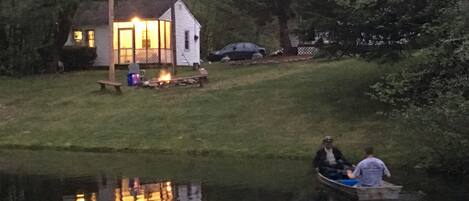 A house with a lit fire on the lawn, a dock, and a person in a boat.