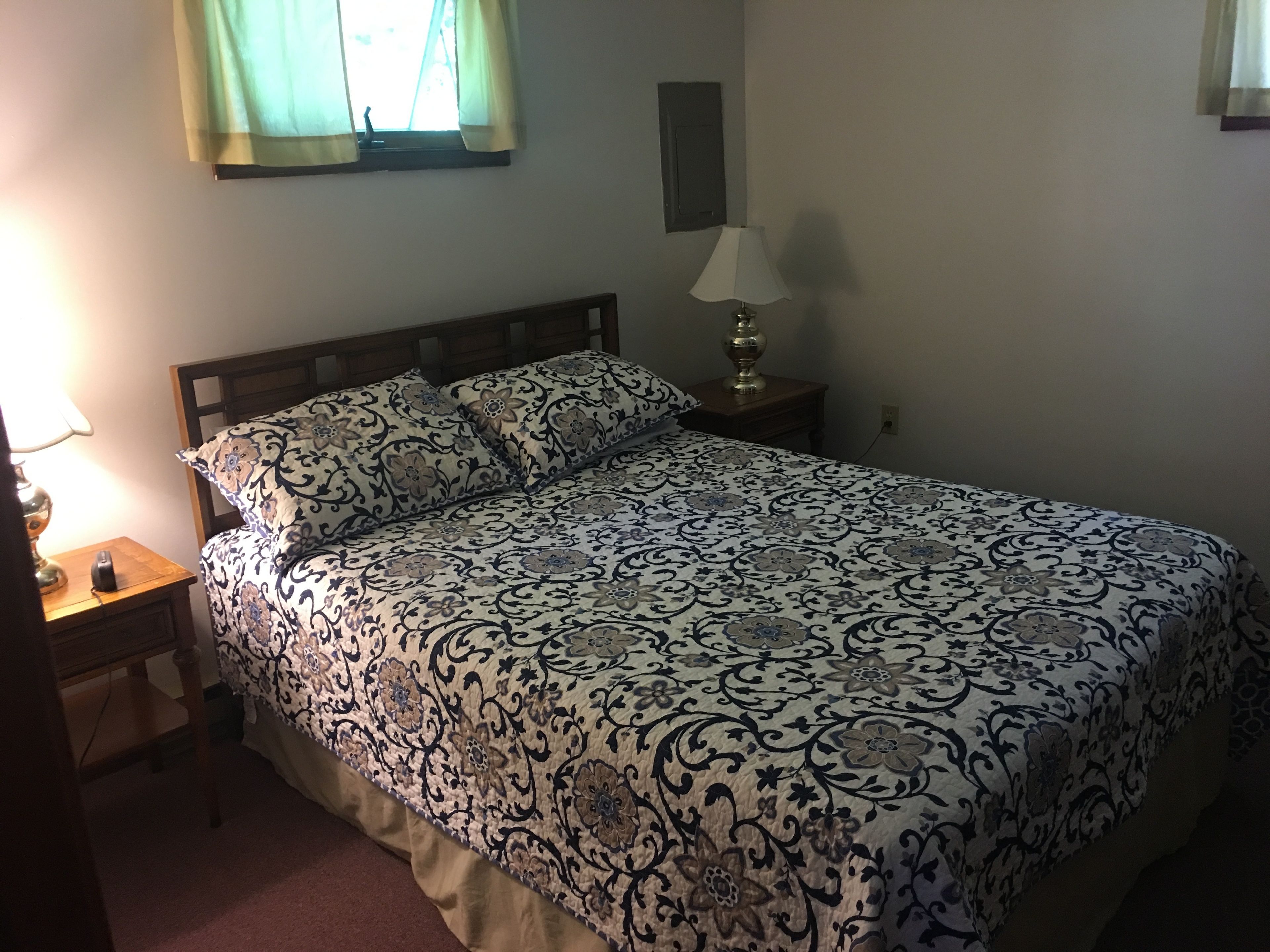 Een slaapkamer met een bed, nachtkastje, lamp en raam met gordijnen.