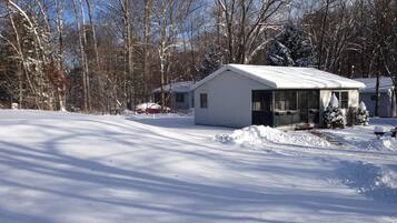 A small, single-story house with a snow-covered roof and a porch, surrounded by leafless trees and a clear sky.