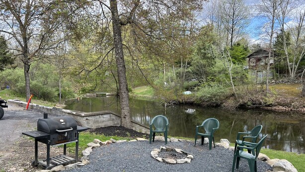 A gravel patio with a fire pit, outdoor seating, and a grill area by a pond. - Echo Valley Cottages (East Stroudsburg)