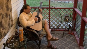 A person sitting on a chair playing a guitar on a porch with a view of a house and yard. - Echo Valley Cottages (East Stroudsburg)