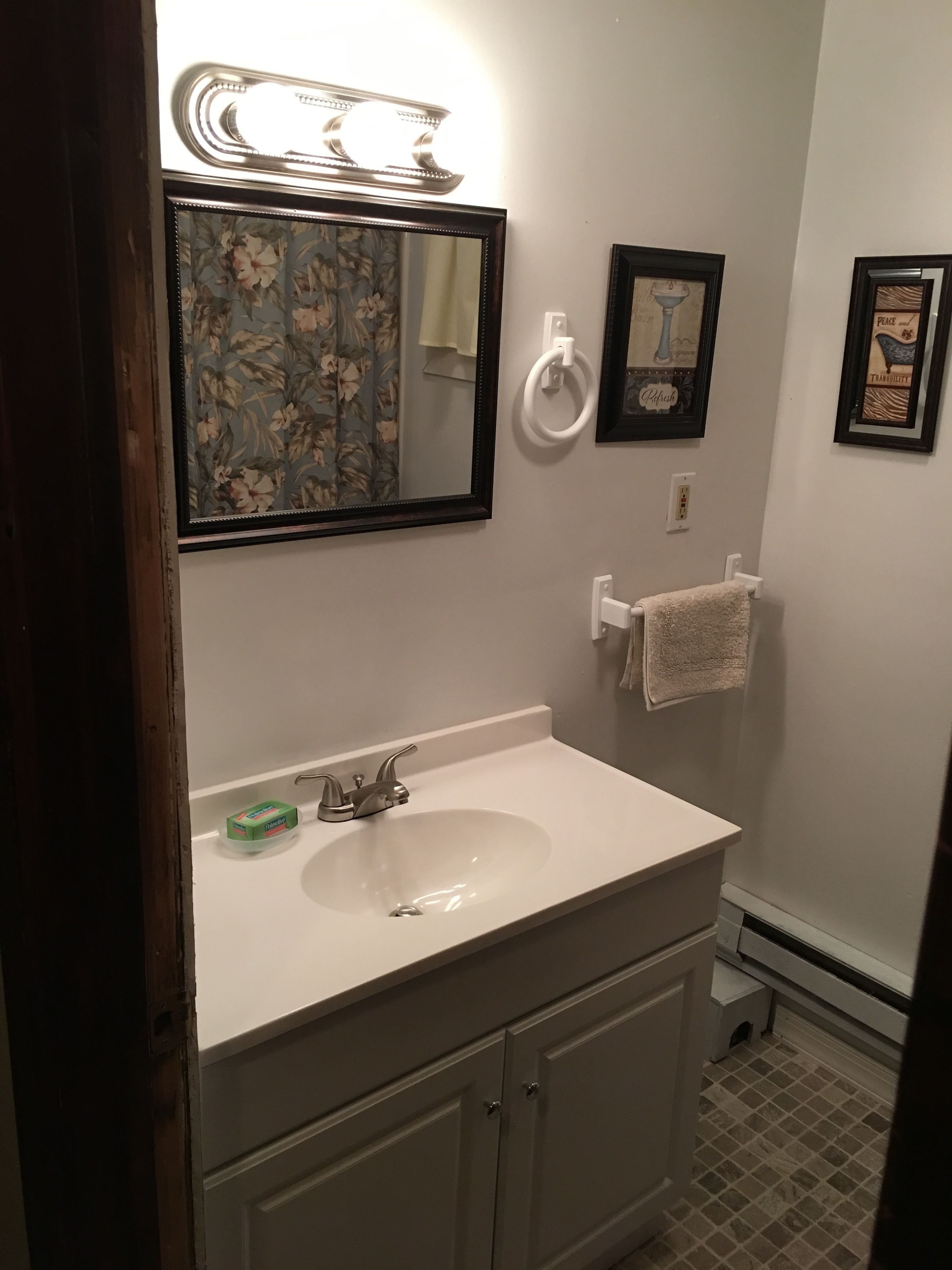 Une salle de bain avec un lavabo blanc, un miroir orné d’un cadre floral, un porte-serviettes et un tableau encadré accroché au mur.