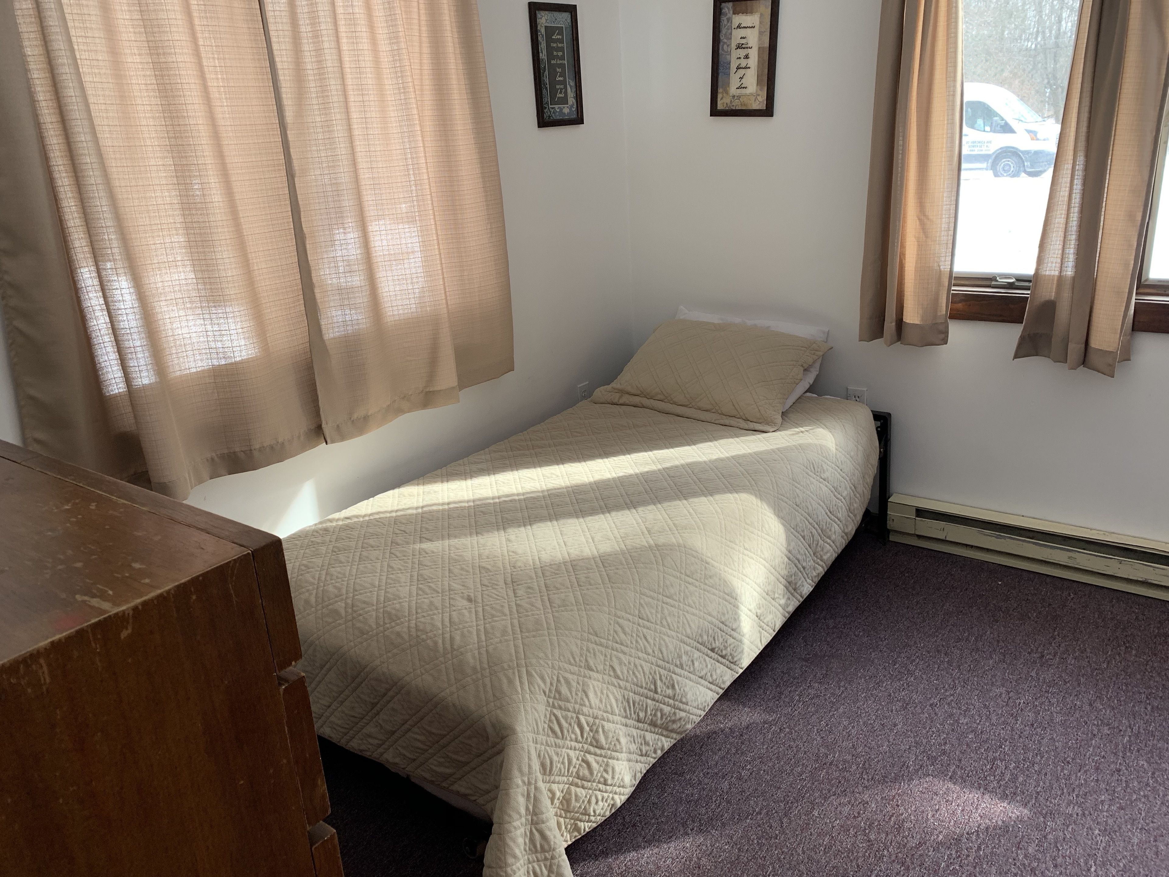 Una habitación pequeña con una cama individual, un escritorio de madera, cortinas color beige y una cita enmarcada en la pared.