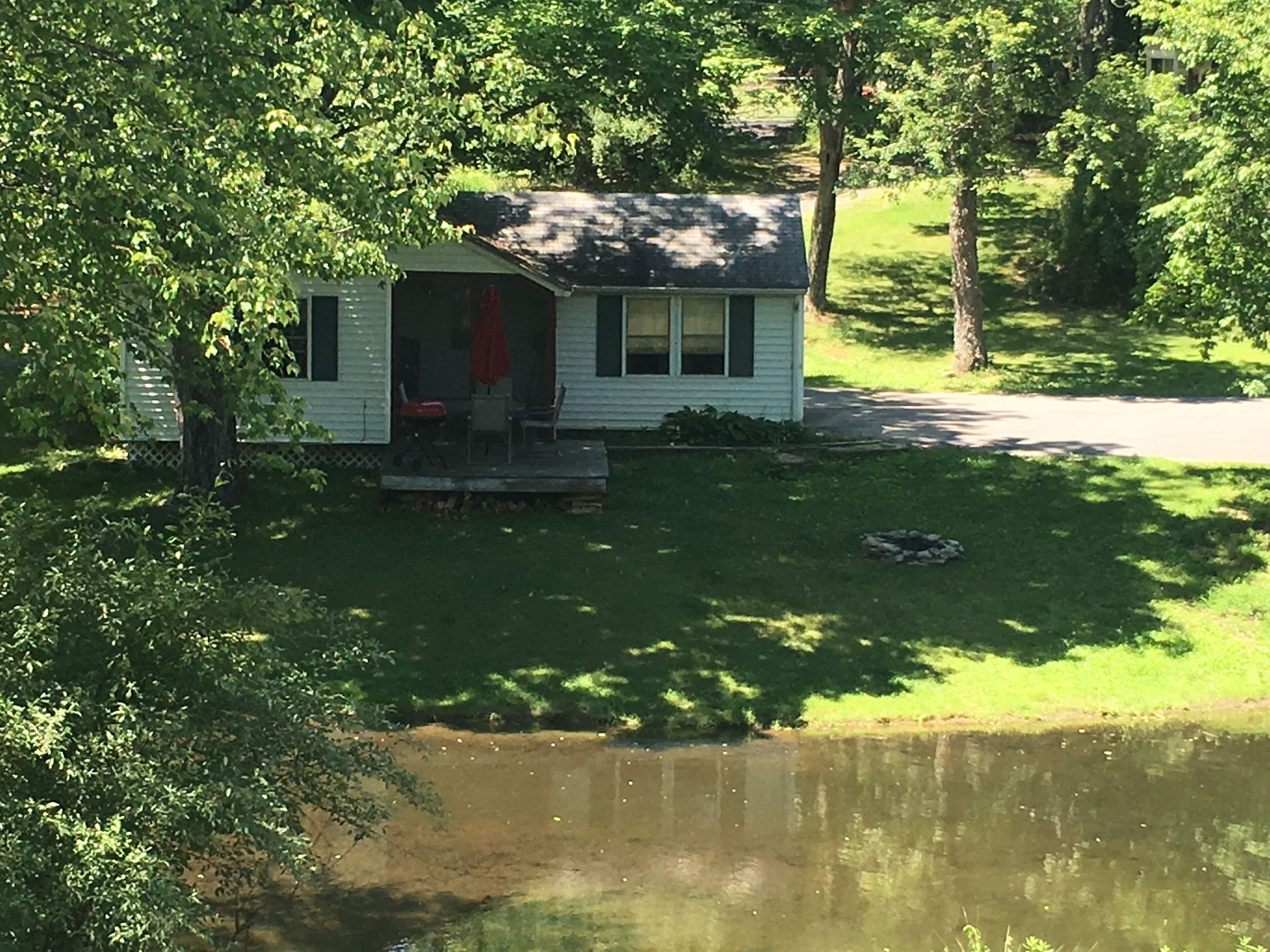A small, single-story house with a porch, situated next to a pond.