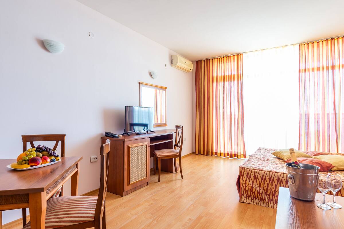 Habitación de hotel con escritorio de madera, una mesa de comedor con fruta, cama con ropa de cama estampada y ventana con cortinas.