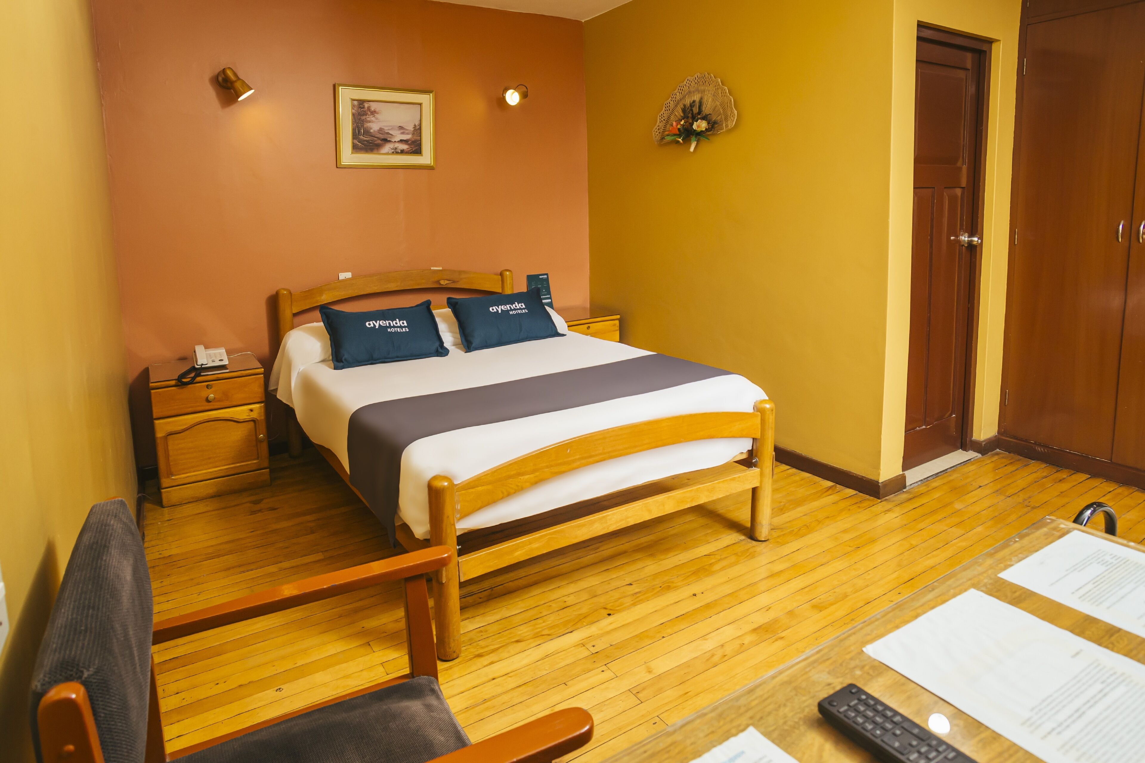 Habitación de hotel con cama de madera, mesita de noche con lámpara, una silla y escritorio con papeles y un control remoto.
