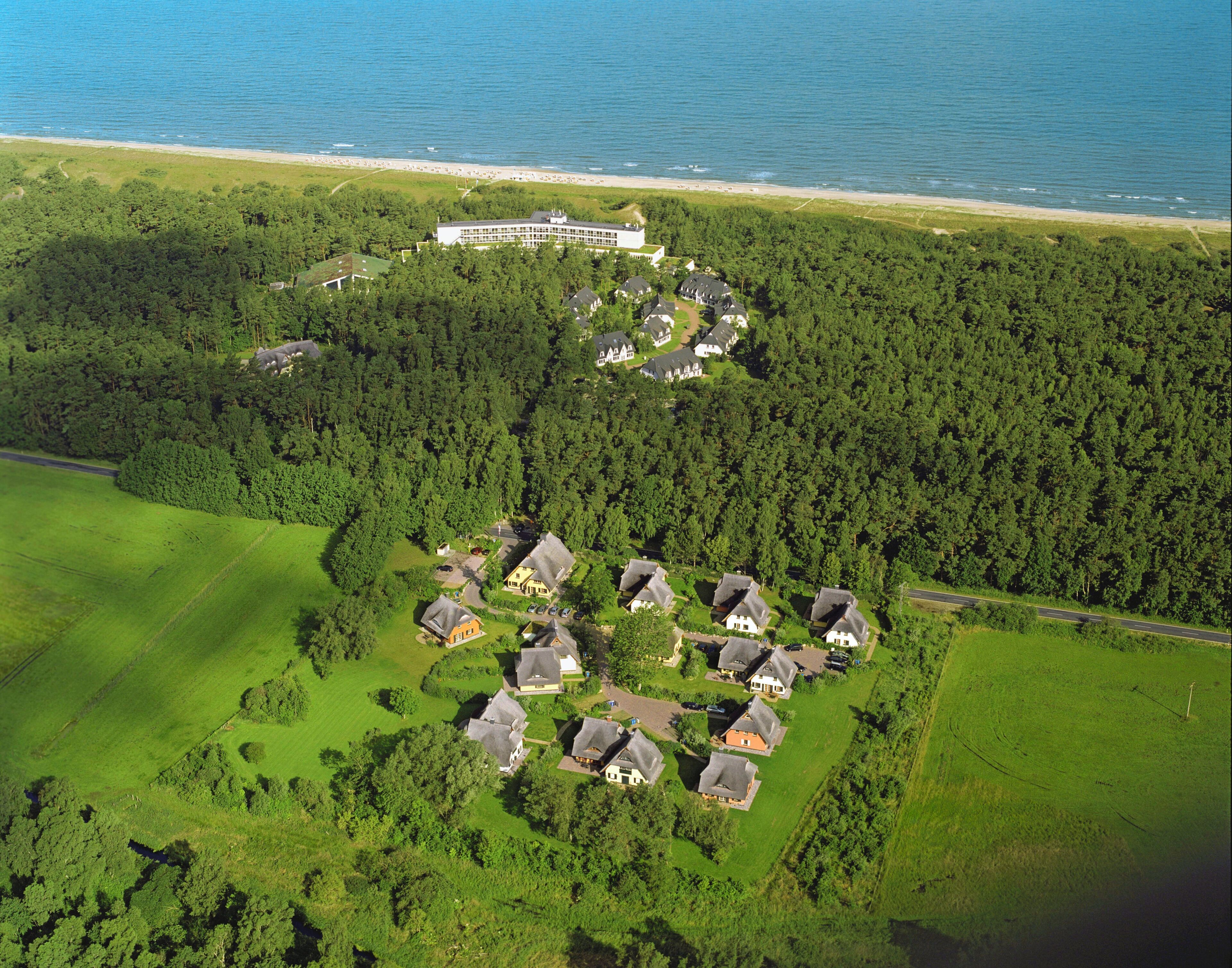 An aerial view of a coastal property with a large building near the beach, surrounded by dense forests and several houses with distinct roofs.