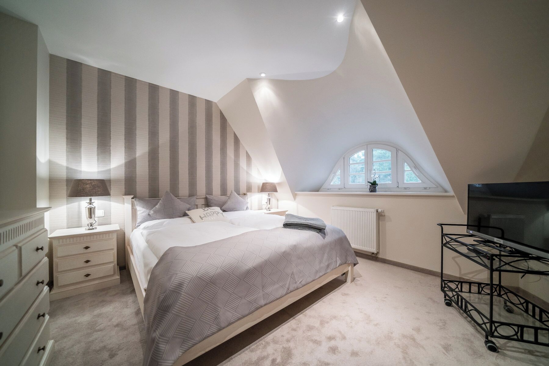 Ein Schlafzimmer mit schrägem Dach, einem Bett mit gemustertem Überwurf, Nachttischlampen, einem Heizkörper, einem Fenster mit Aussicht und einem Fernseher auf einem Schrank.
