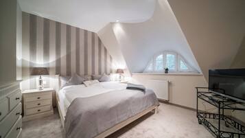 Ein Schlafzimmer mit schrägem Dach, einem Bett mit gemustertem Überwurf, Nachttischlampen, einem Heizkörper, einem Fenster mit Aussicht und einem Fernseher auf einem Schrank.