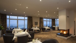 Ein Restaurant mit gedeckten Tischen, einem Kamin und großen Fenstern mit Meerblick.