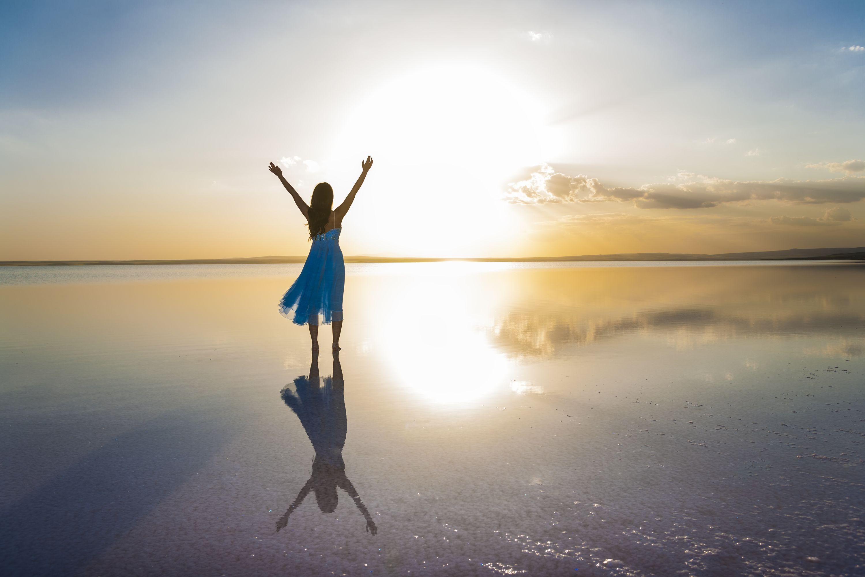 A person in a blue dress standing on a reflective surface with arms raised, facing a sunset.
