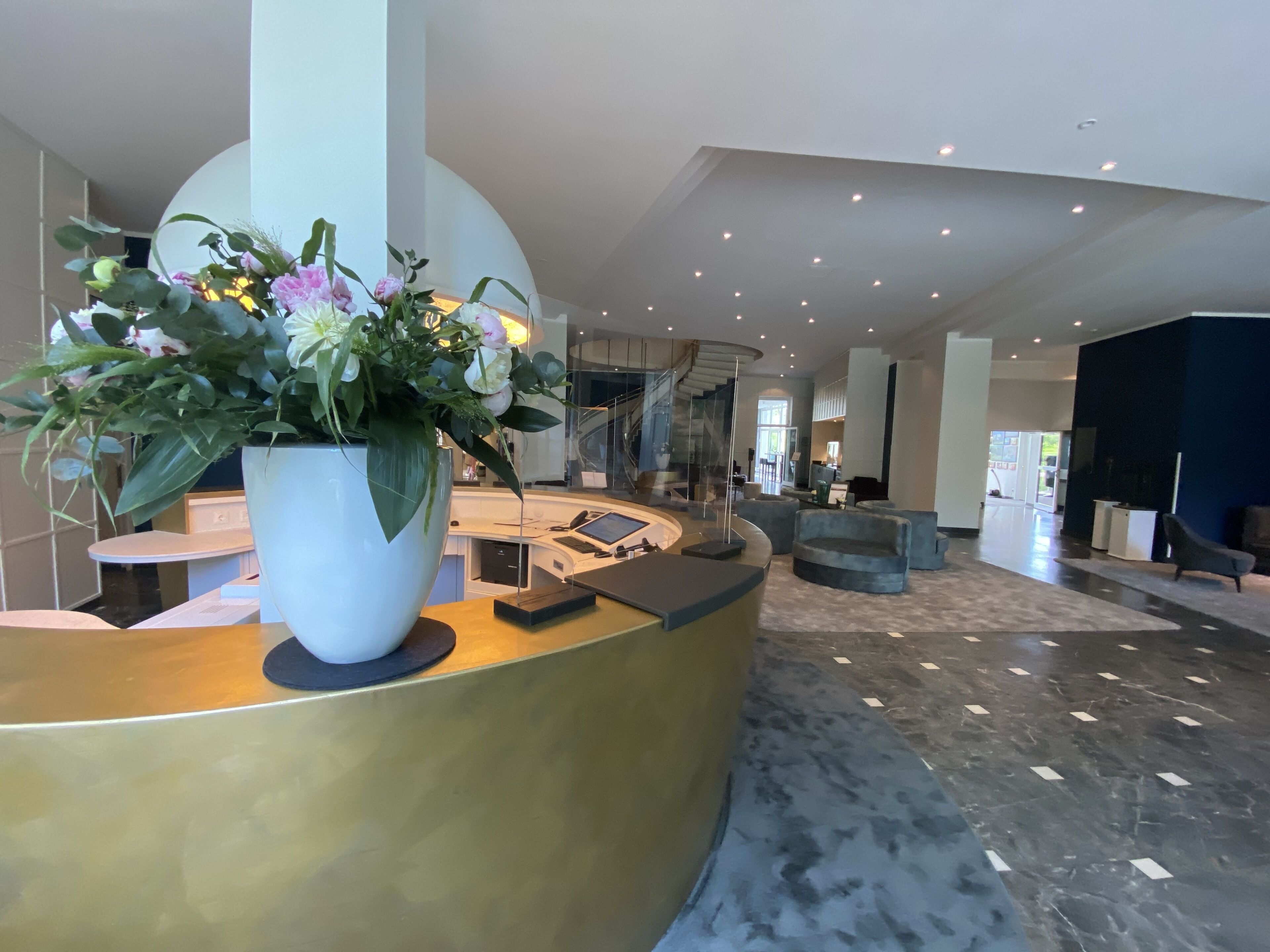 Un espace d’accueil moderne avec un comptoir incurvé doré, un grand vase blanc rempli de fleurs et un escalier en arrière-plan.