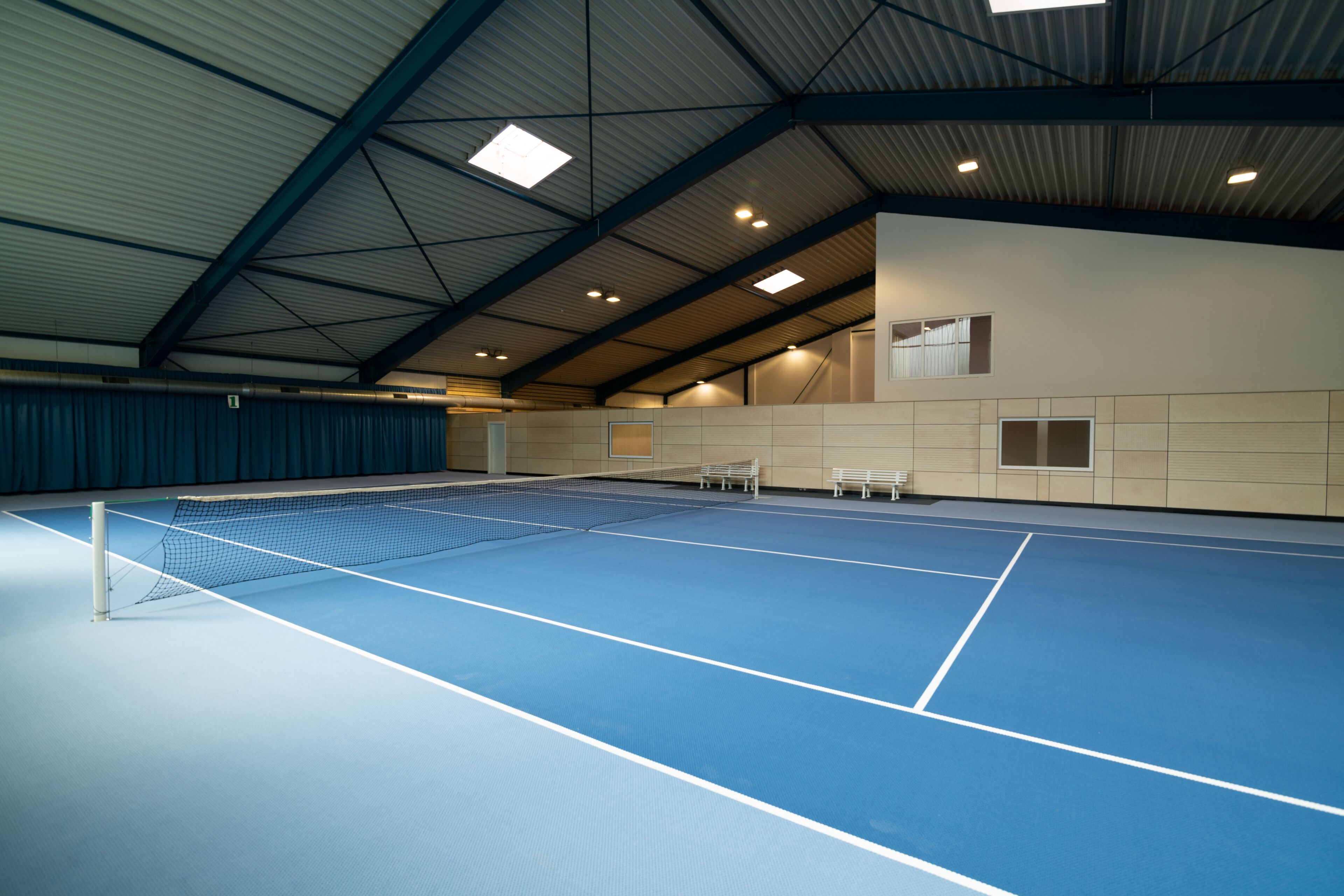 Un court de tennis intérieur doté d’une surface de jeu bleue, d’un filet blanc et de bancs le long des murs.