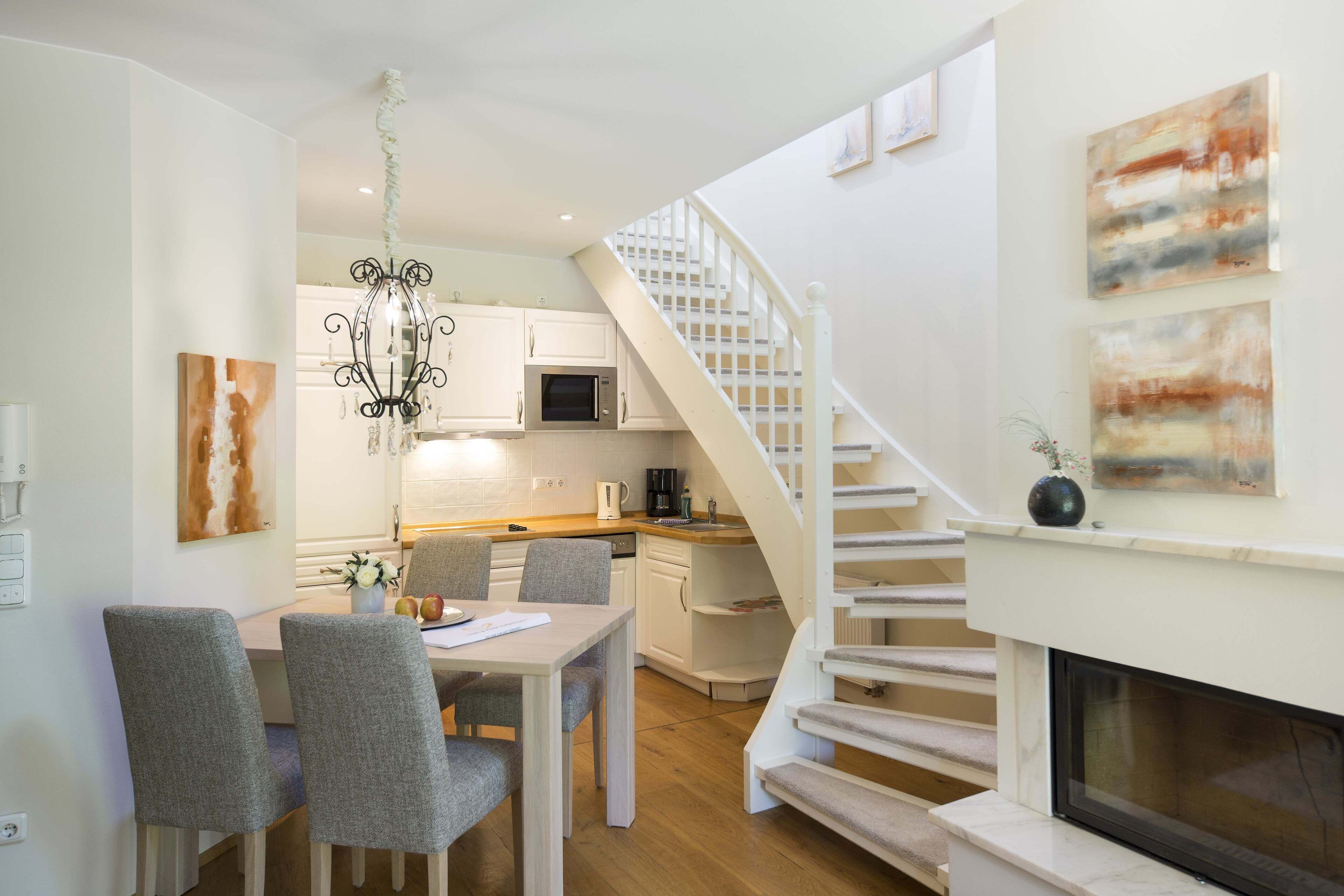 A modern kitchen with white cabinets, a microwave, and a coffee maker. There is a dining area with a table and chairs. A staircase with a white railing leads to a living area with a fireplace.