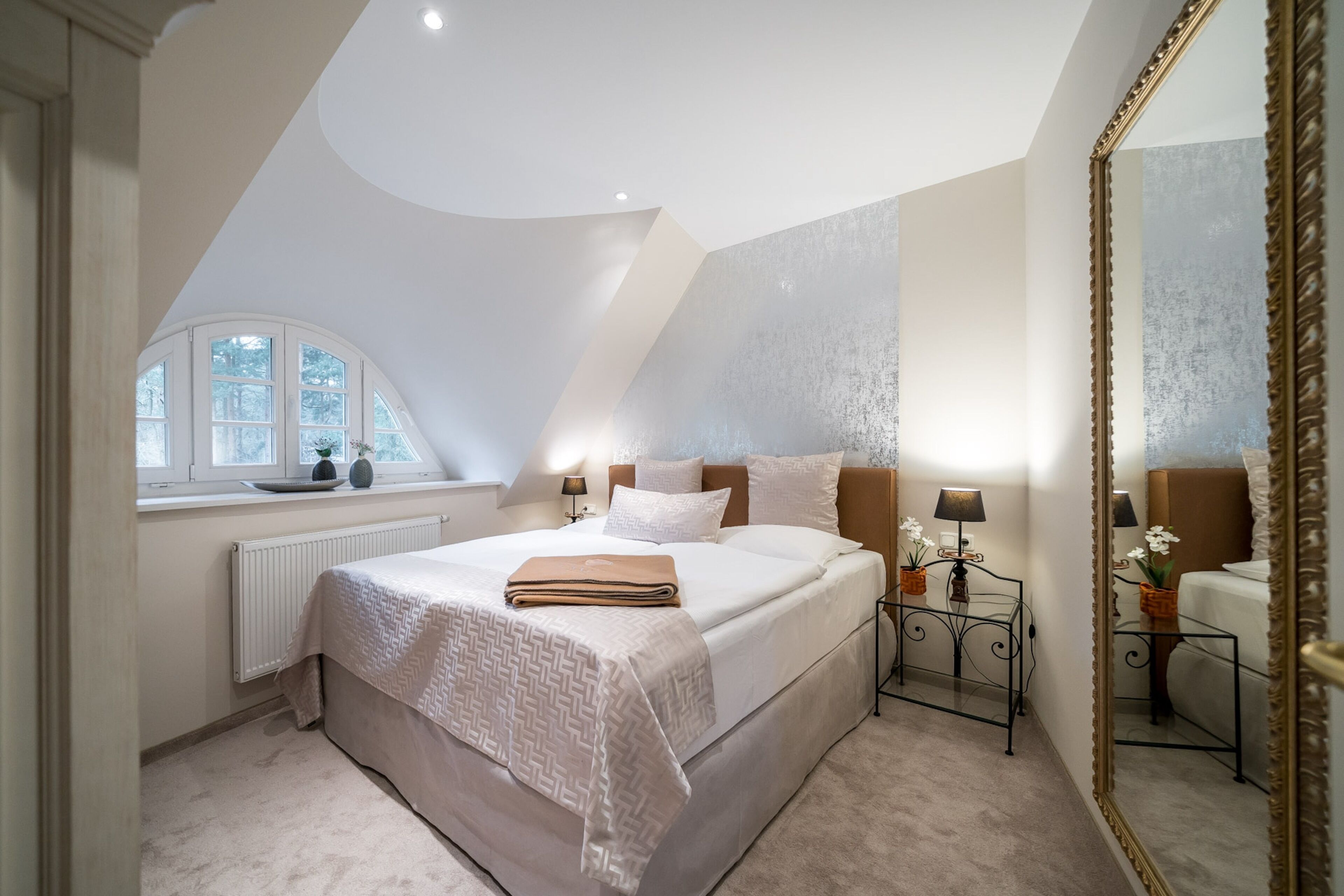 A bedroom with a large bed, a window with a view, a bedside table with lamps, and a mirror.
