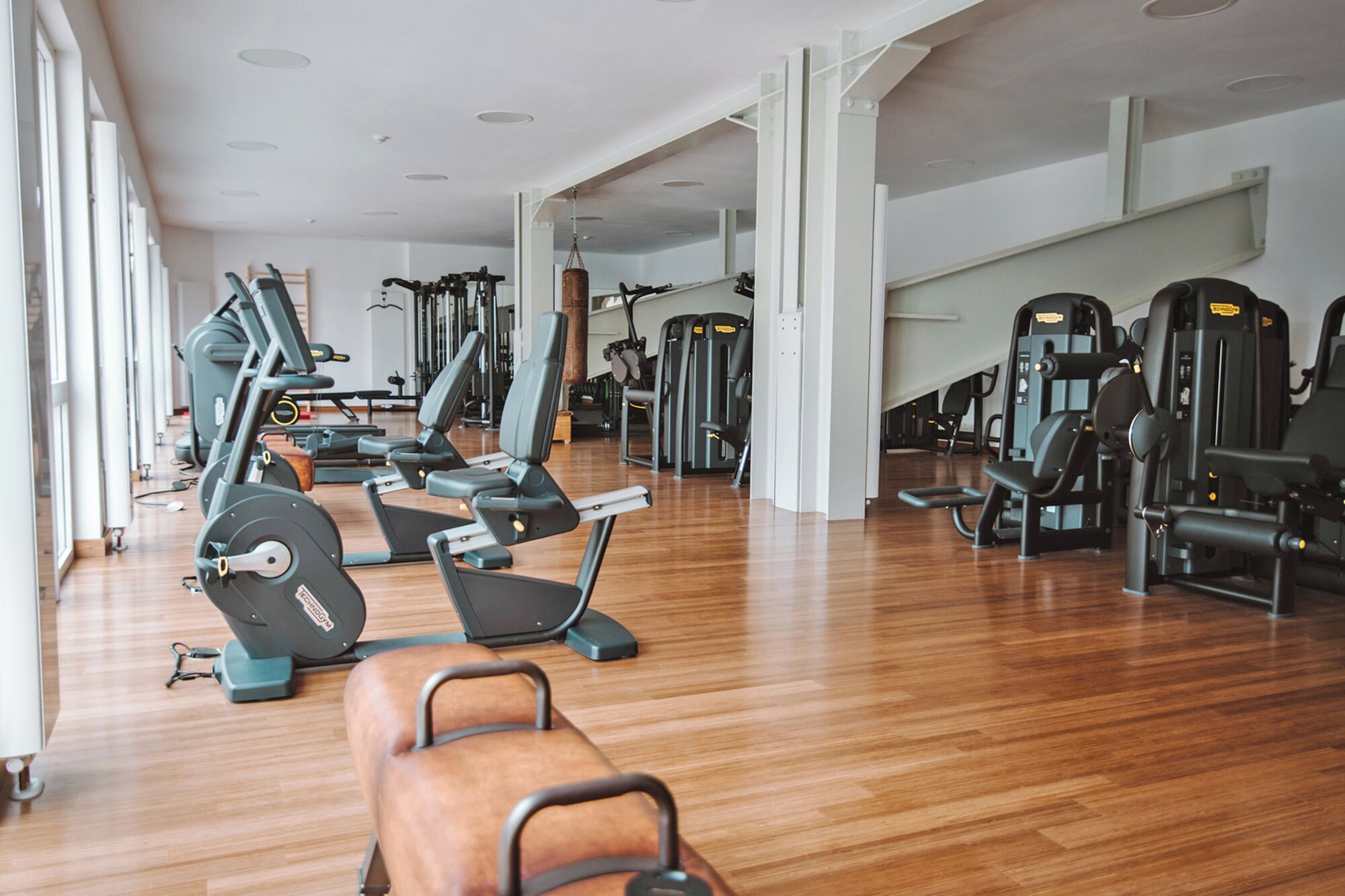 Une salle de sport spacieuse, dotée de divers appareils d’exercice, d’un plancher en bois et de grandes fenêtres qui laissent entrer la lumière naturelle.
