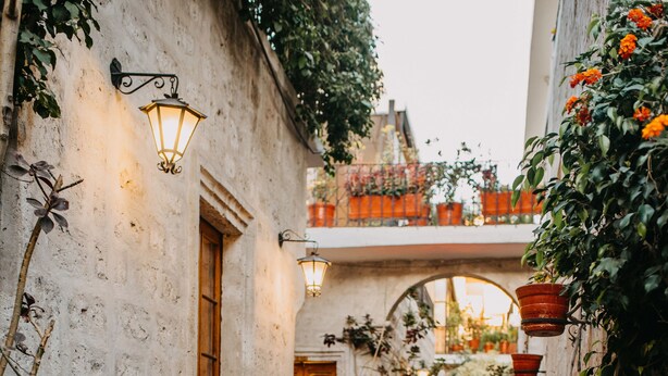 Une ruelle étroite où l’on voit une femme tirant une valise, une fontaine et des plantes en pot.