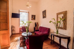 Una sala de estar con un sofá rojo, una mesa de centro de vidrio y suelo de madera. Se observa una ventana con barrotes y una araña.