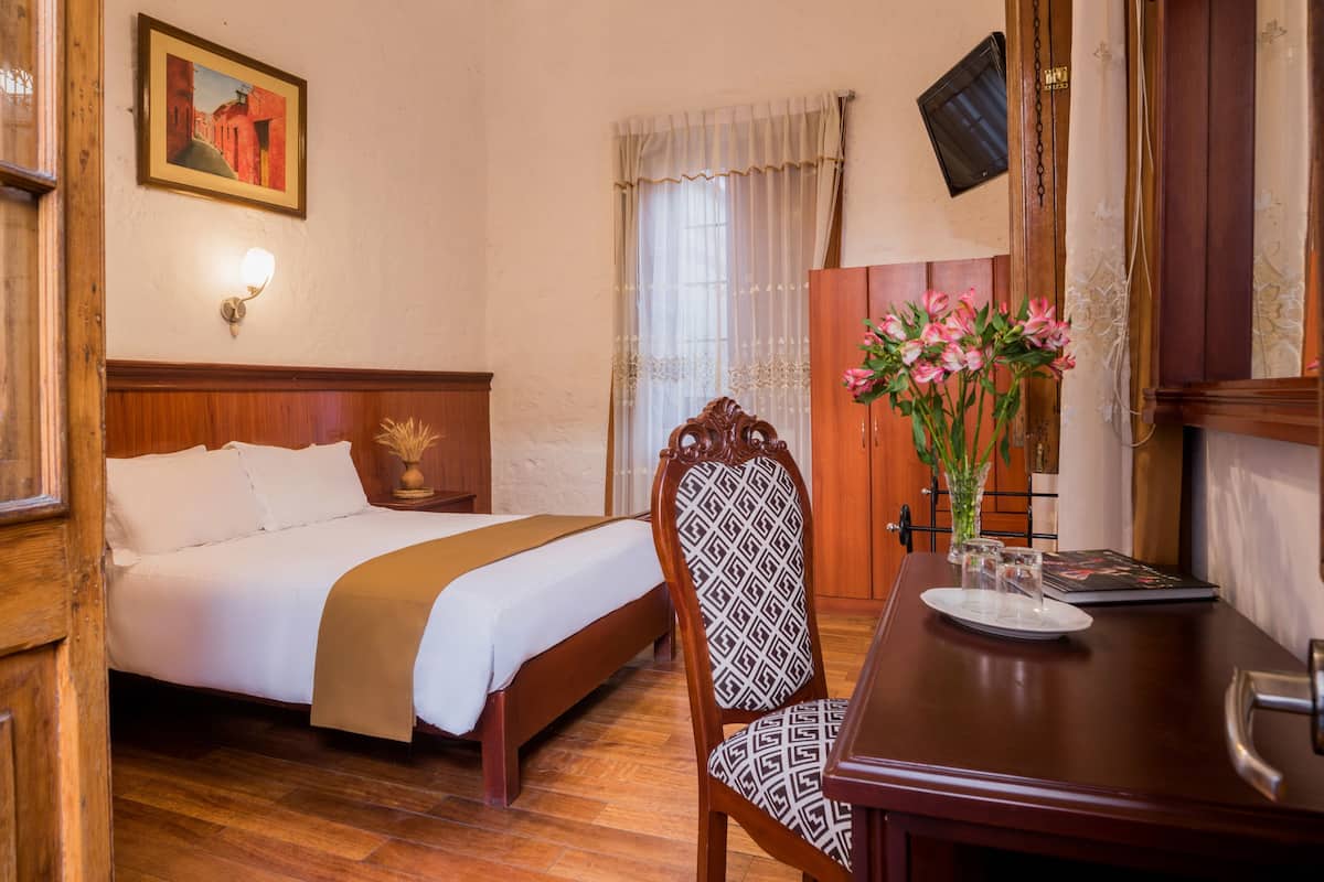 Una habitación de hotel con una cama, un escritorio de madera, una silla, un jarrón con flores, un televisor montado en la pared y un cuadro en la pared.