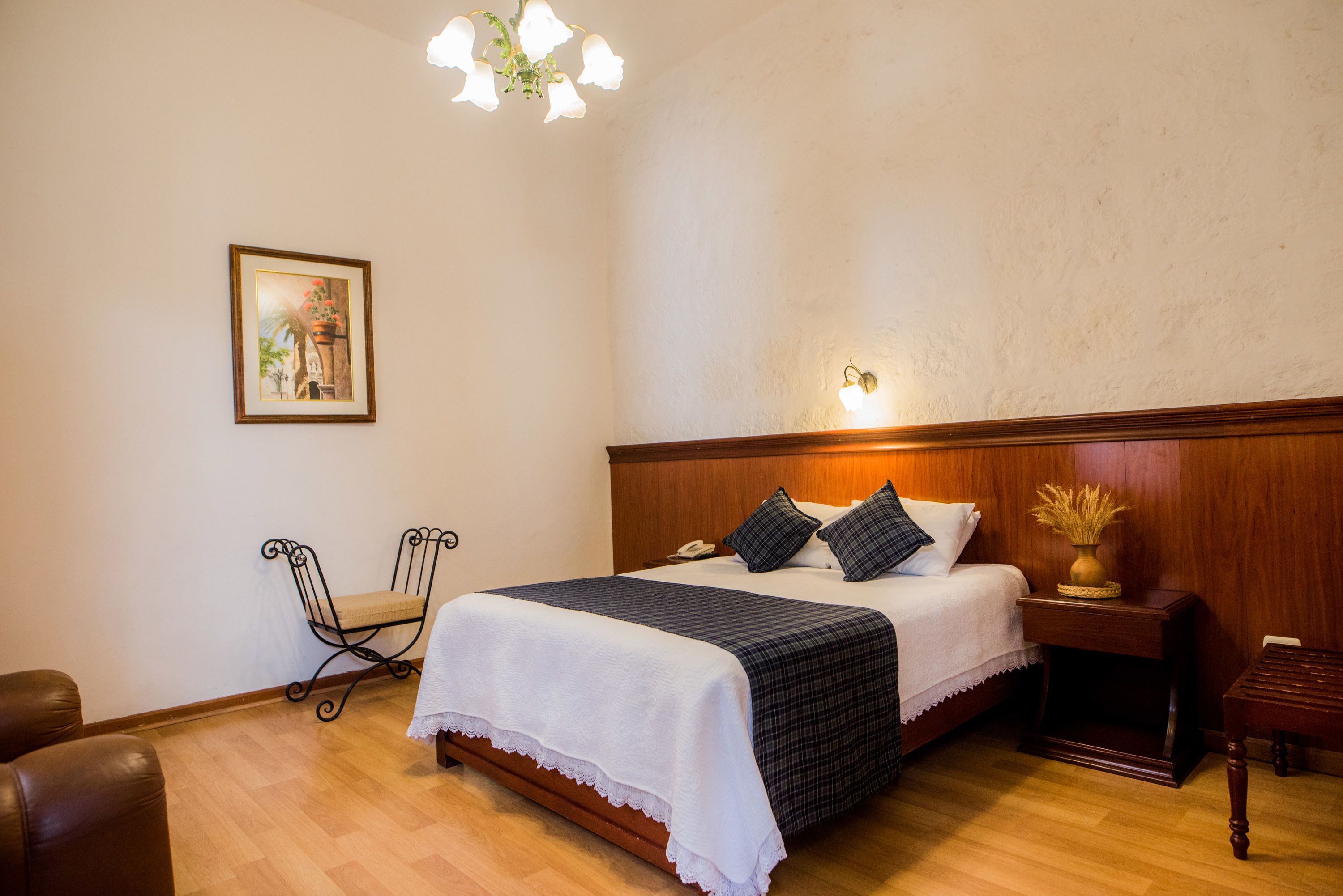 A bedroom with a wooden headboard, a bed with a white and dark checkered bedspread, two pillows, a bedside table with a vase, and a framed picture on the wall.