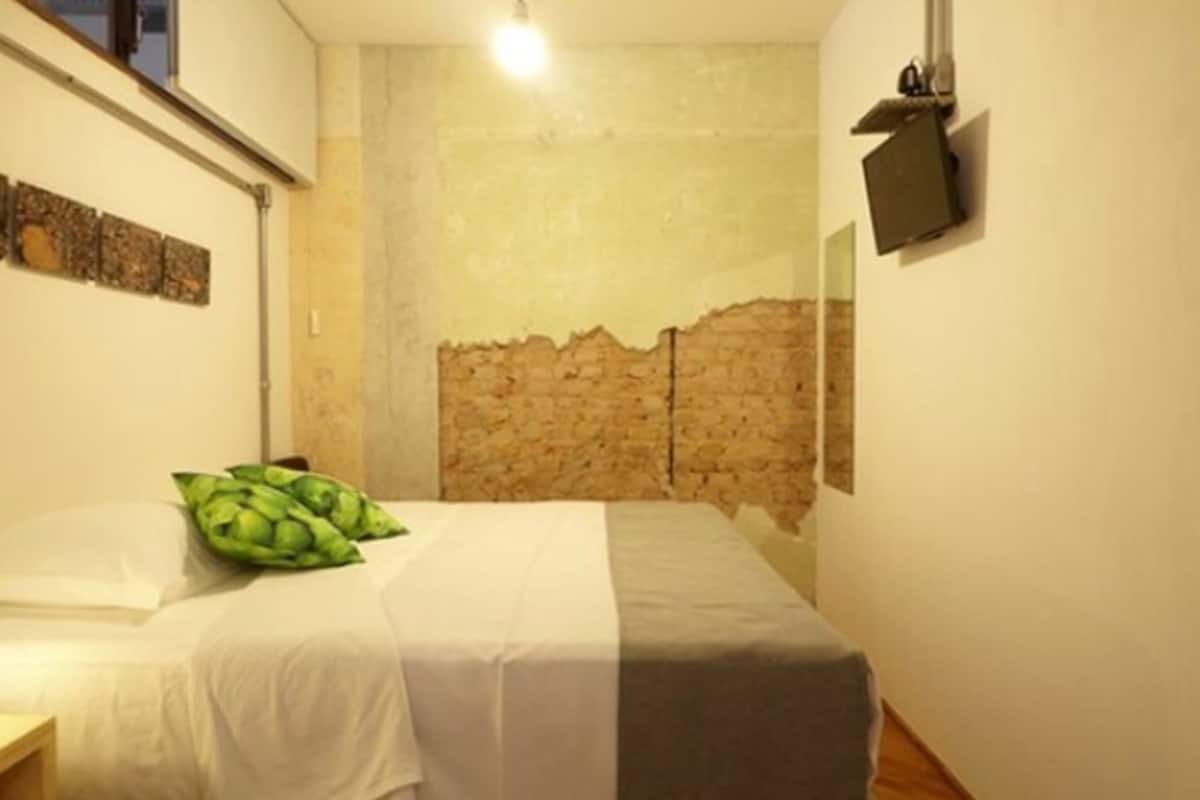 A small, narrow room with a bed, a stone wall, a mounted TV, and a small table.
