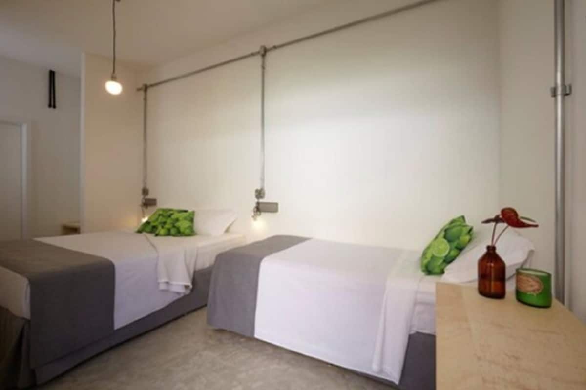 A room with two single beds, a small table with a bottle and a container, and a hanging light fixture.