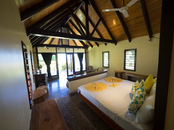 A bedroom with a large bed, wooden ceiling beams, a window with curtains, and a view of the outdoors.