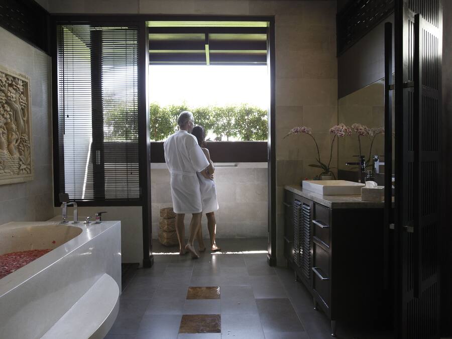 Un baño moderno con una bañera grande, un lavamanos y un espejo. Se ve una puerta que da a un balcón con vista a árboles.