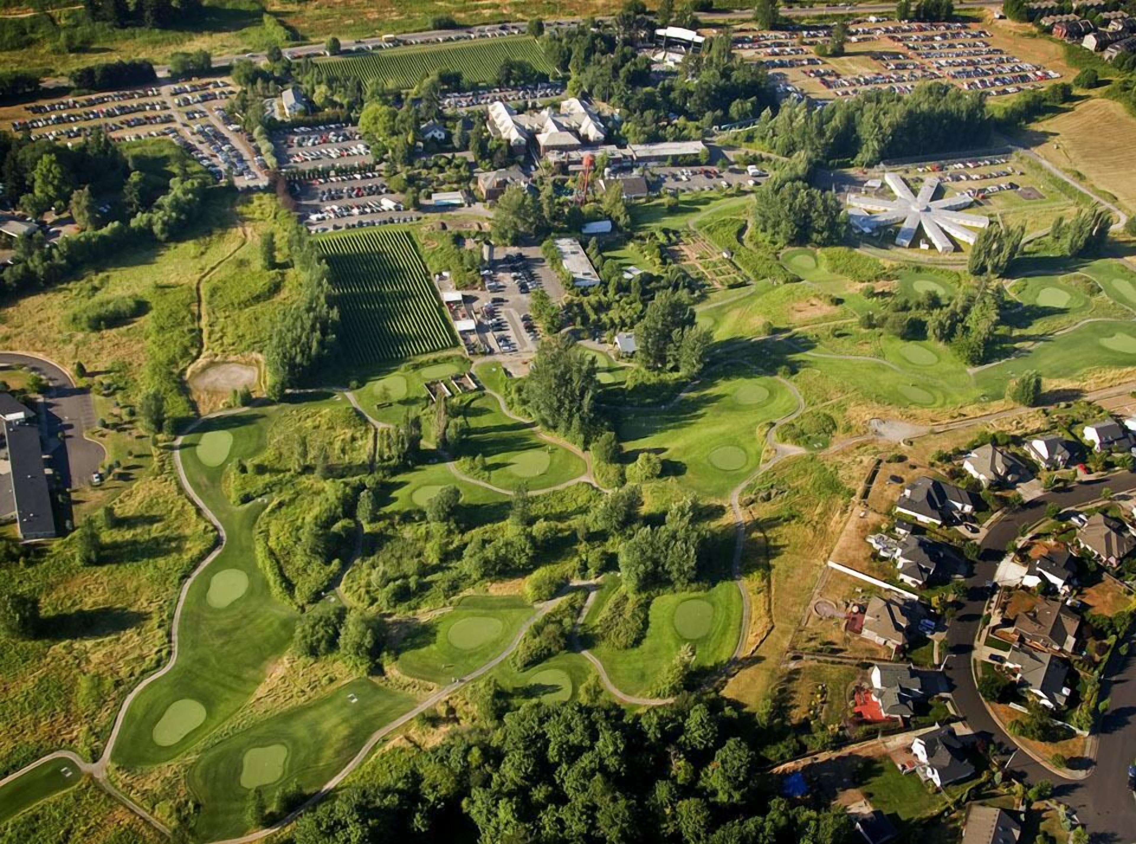 An aerial view of a golf course with multiple greens, a large parking area, and residential buildings.