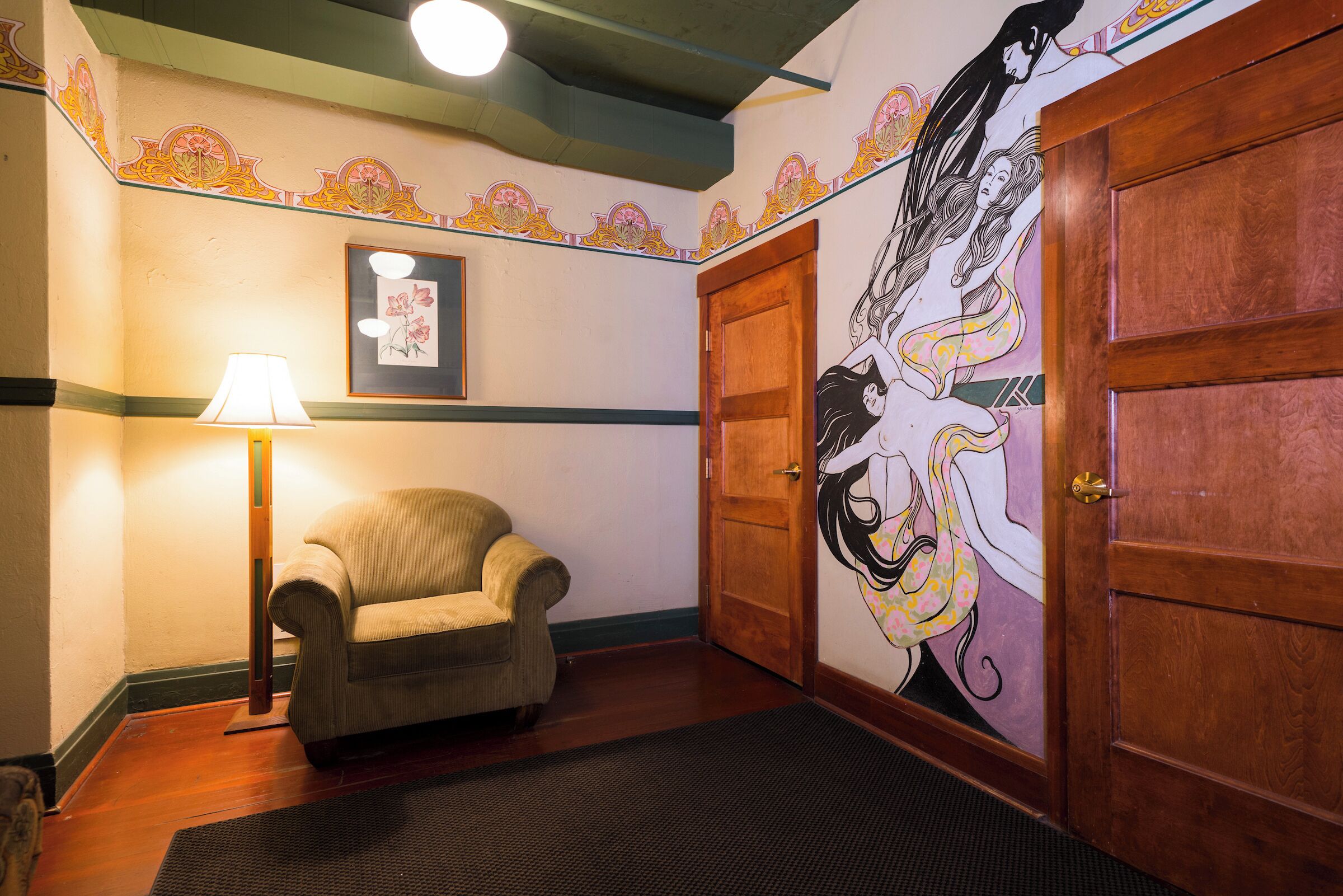 A hallway with a wooden door, a framed picture on the wall, a lamp, and a chair.