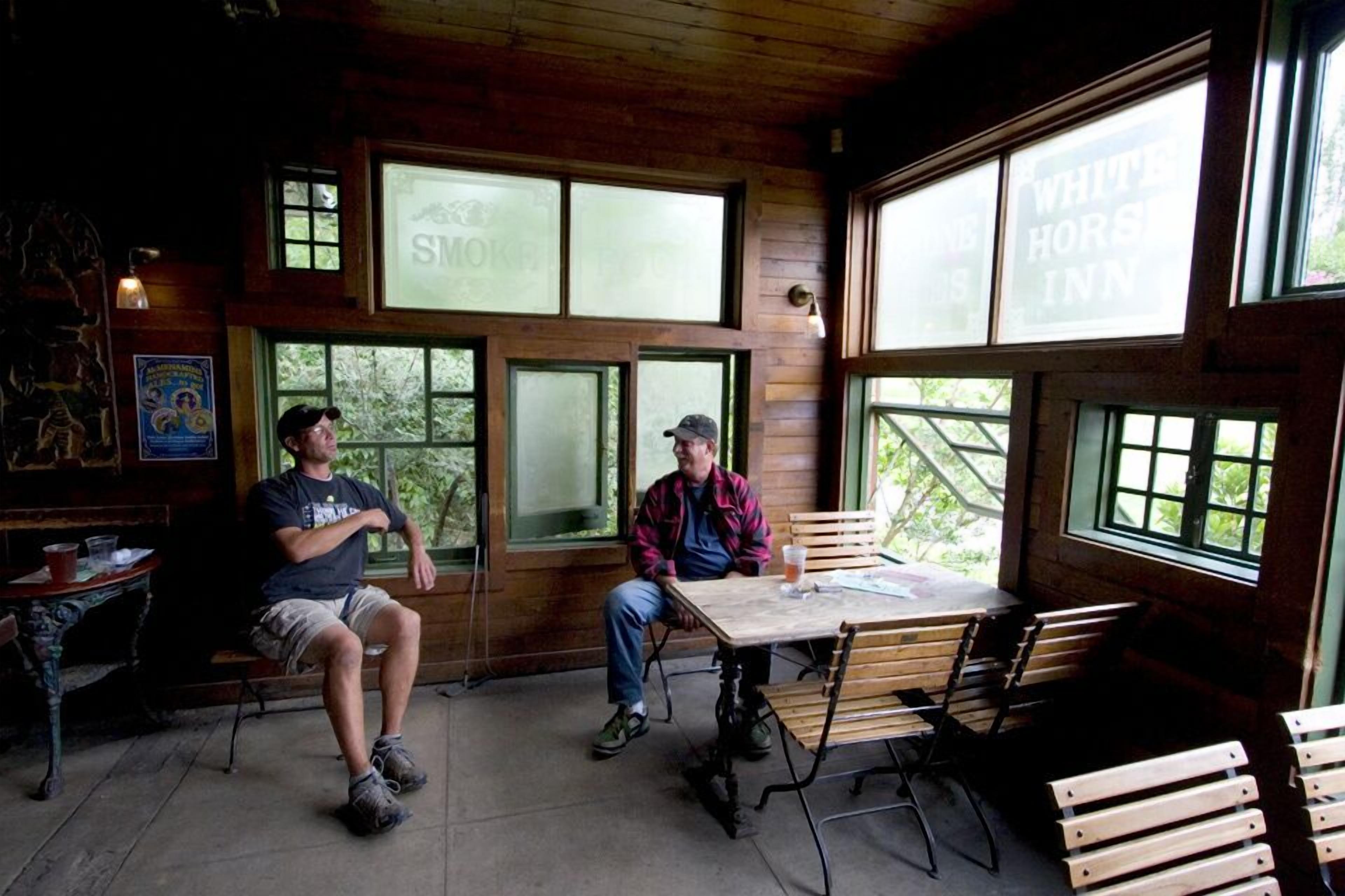 A rustic wooden interior with two men seated at a table, large windows, and a sign reading "WHITE HORSE INN".