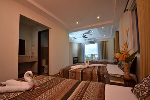 Quarto de hotel com duas camas, ventoinha no teto, vista para o mar e área de refeições com mesa e cadeiras.