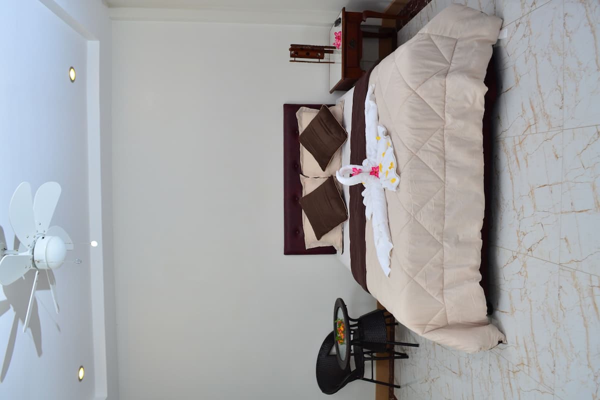A room with a bed, a small table with a chair, a ceiling fan, and a marble-patterned floor.