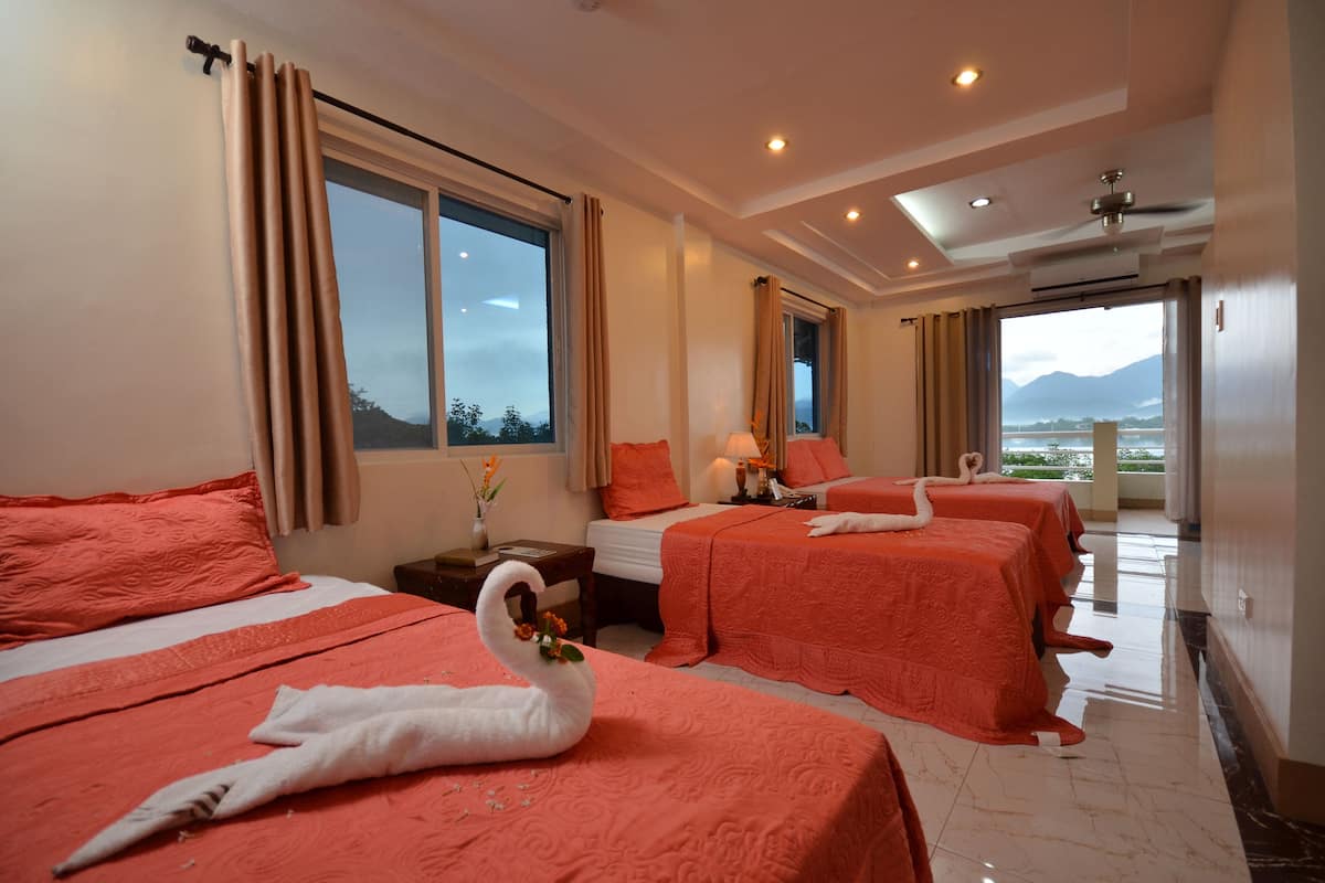 A hotel room with three beds, each with a swan decoration, a large window with curtains, a bedside table with a lamp, and a view of mountains.