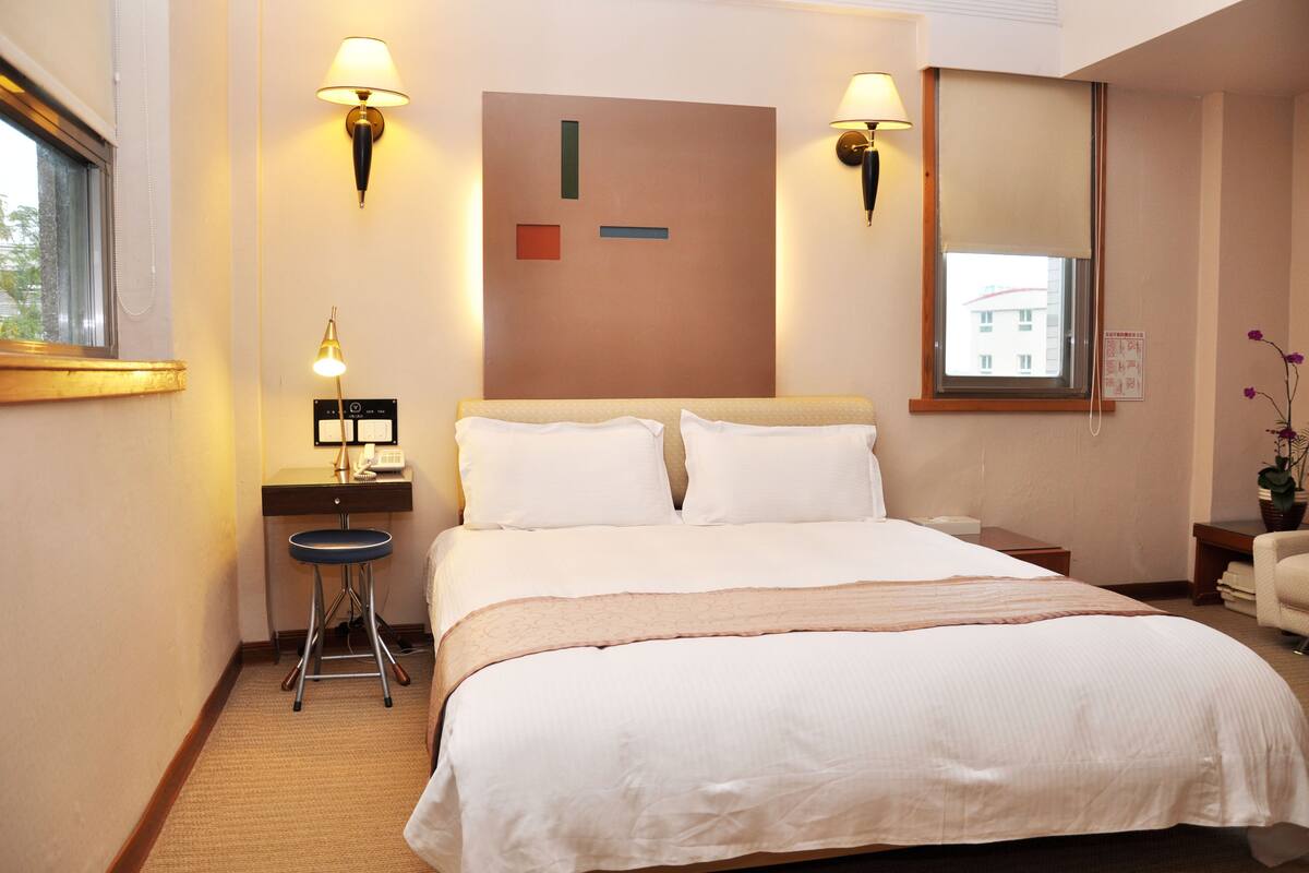A hotel room with a bed, bedside lamps, a desk with a lamp, a chair, and a window.