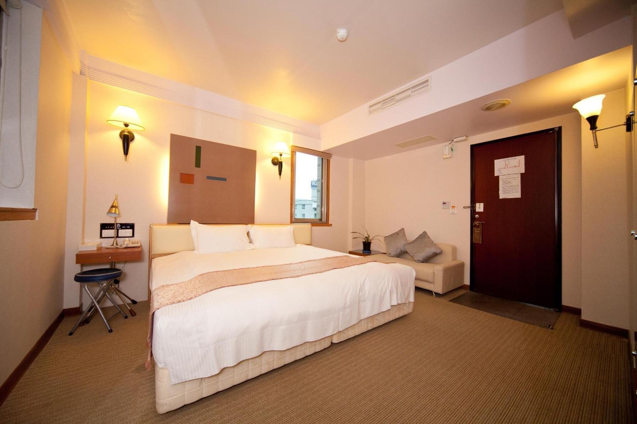 A hotel room with a large bed, bedside lamps, a desk with a telephone, and a door with a sign.