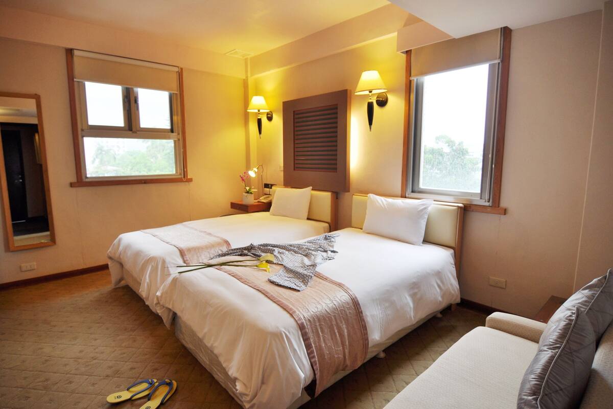 A hotel room with two beds, a sitting area, a window, and a lamp.