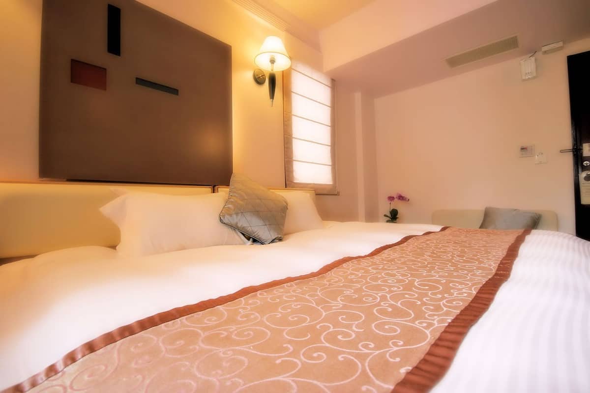 A hotel room with a bed, a nightstand, a lamp, a window, and a wall-mounted clock.