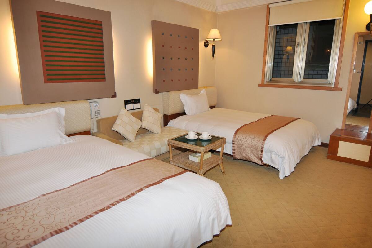 A hotel room with two beds, a bedside table with cups, a wall-mounted control panel, and a window with a view.